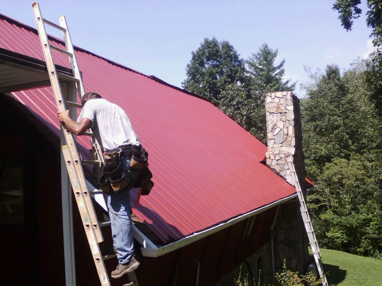 Roofer on a ladder beside a red metal roof and stone chimney — Soffit & Fascia Repair by Roofing Brothers in Castaic